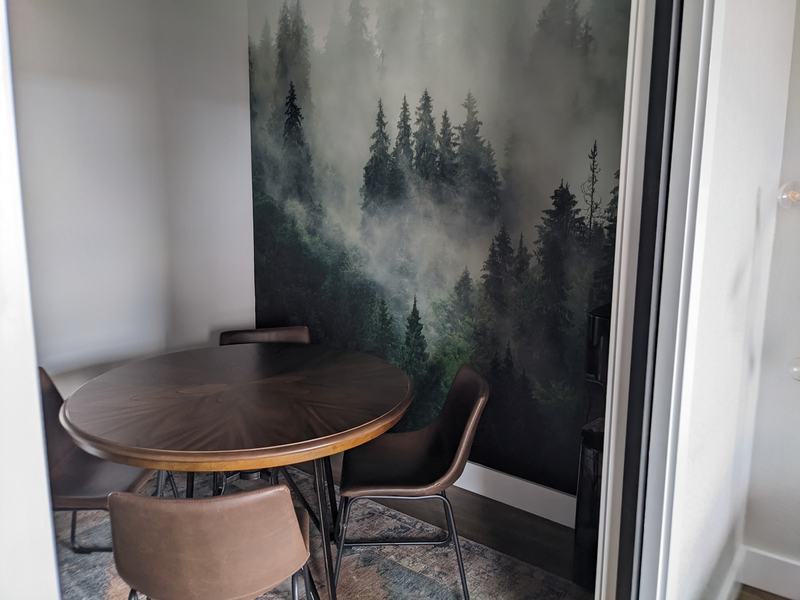 A small breakout nook with a misty evergreen forest wall mural, a round wood table, and brown leather chairs.